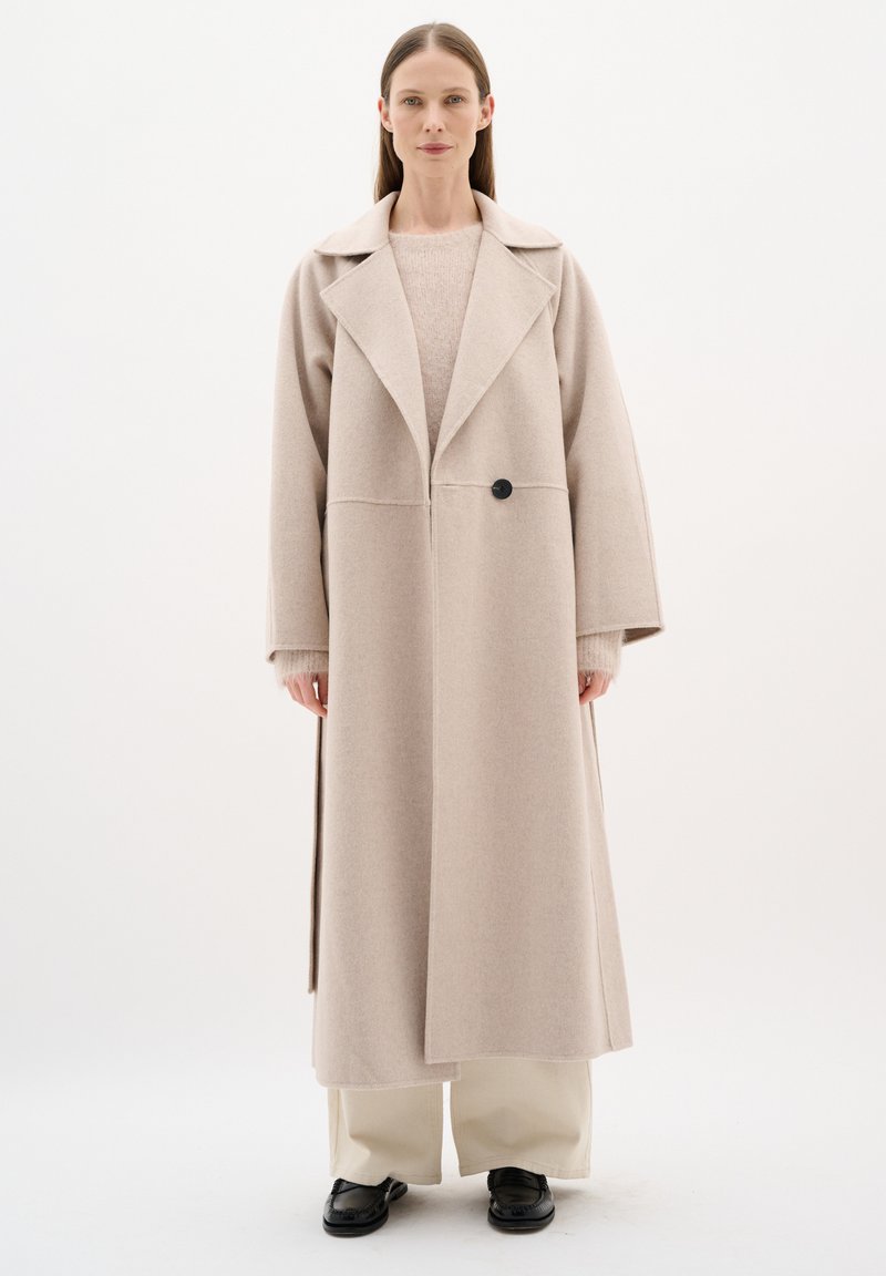 Cappotto in misto lana beige con ampi rever, maniche lunghe e un unico bottone nero. Ha una vestibilità dritta e scende sotto il ginocchio.