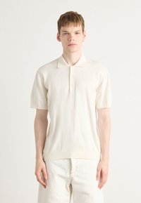 Polo crème à manches courtes, texture tricotée, col doux, patte de boutonnage à trois boutons et ourlet côtelé. Associé à un pantalon de couleur claire.