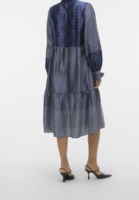 Vestito blu navy con maniche lunghe, strati arricciati e vestibilità comoda. Il tessuto ha una texture liscia con diagrammi geometrici.
