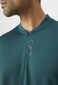 Zoomiran posnetek osebe, ki nosi teal strukturno majico z zadrgo in logotipom "EA7 Emporio Armani" na prsih ter gladkim ovratnikom.