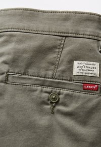 Tela verde oliva con una textura suave, que presenta un bolsillo de parche, cierre de botón y una etiqueta de Levi's con acento rojo.