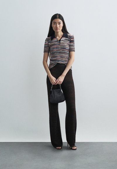 Woman wearing a striped short-sleeve top, sheer black patterned pants, holding a small textured handbag against a plain light background.