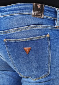 Los jeans de mezclilla azul presentan una etiqueta de cuero triangular en el bolsillo trasero con "GUESS" grabado. La costura es de color amarillo, resaltando el diseño.