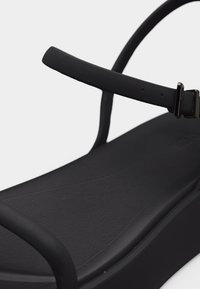 Μαύρο σανδάλι πλατφόρμα με λεπτές ραβδώσεις και κούμπωμα με πόρπη πάνω σε υφασμένο πέλμα.