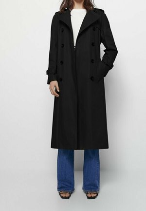Femme portant un long trench-coat noir croisé sur un haut blanc, un jean bleu et des talons noirs ouverts à l’avant, debout avec une main dans la poche.