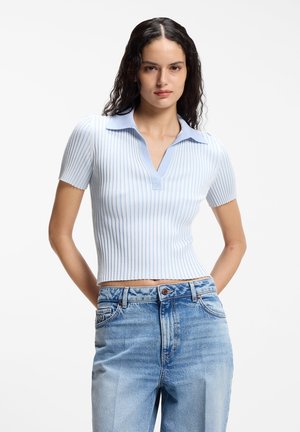 Kobieta o długich kręconych włosach, ubrana w jasnoniebieski sweter w paski z krótkim rękawem i wyblakłe dżinsy z wysokim stanem, stojąca z rękami za plecami.