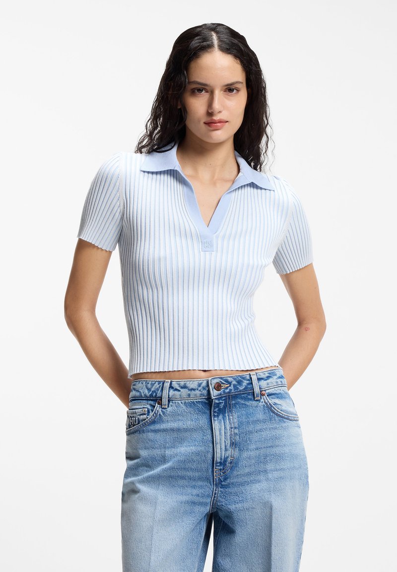 Γυναίκα με μακριά σγουρά μαλλιά που φοράει πλεκτό τοπ με κοντά μανίκια σε ανοιχτό μπλε ριγέ και ψηλόμεσο ξεθωριασμένο τζιν, στέκεται με τα χέρια πίσω από την πλάτη.