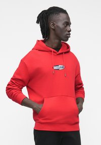 Κόκκινη βαμβακερή μπλούζα με κουκούλα, μπροστινή τσέπη και ρυθμιζόμενο κορδόνι. Διαθέτει λογότυπο "GUESS JEANS" στο στήθος. Λεπτή υφή.