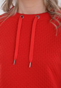 Rode hoodie met textuur, voorzien van een ronde halslijn en zilverkleurige koordjes. De stof heeft een subtiel patroon, wat de visuele interesse vergroot.