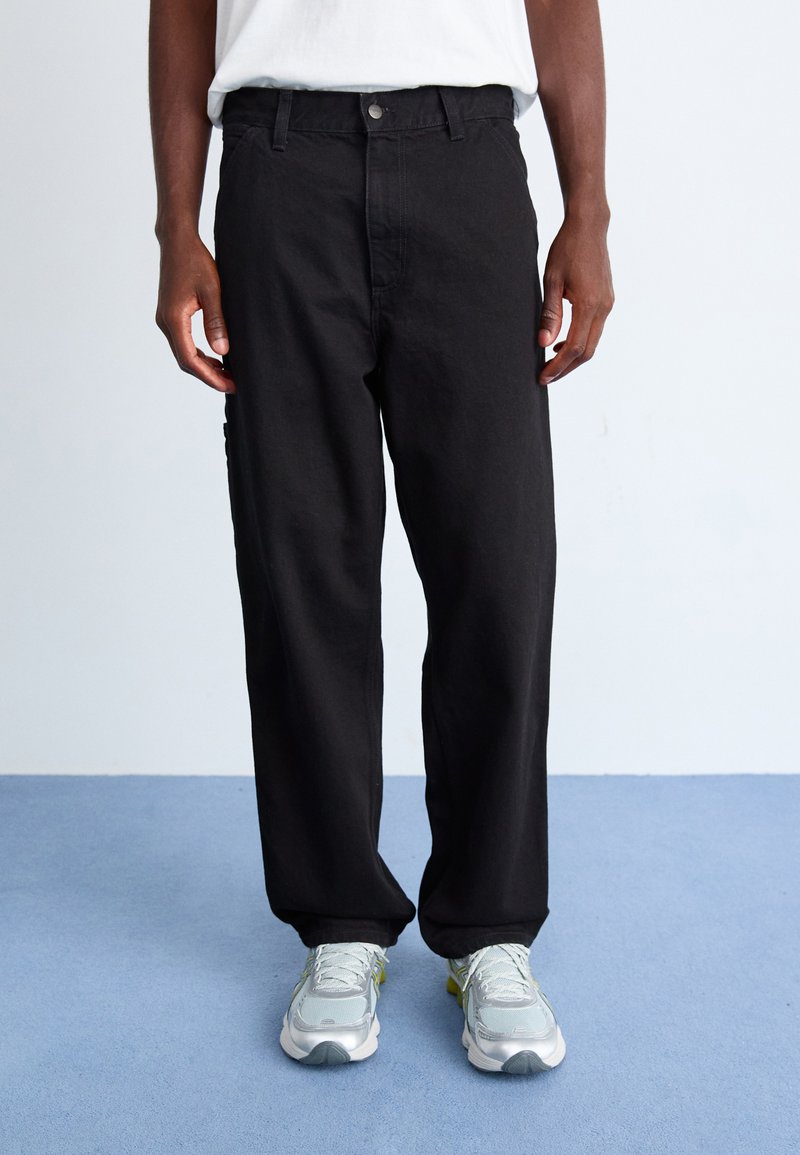 Pantalons en denim noirs, larges et confortables, avec une fermeture à bouton et à glissière, dotés de poches latérales et d'un design épuré, associés à des baskets grises.