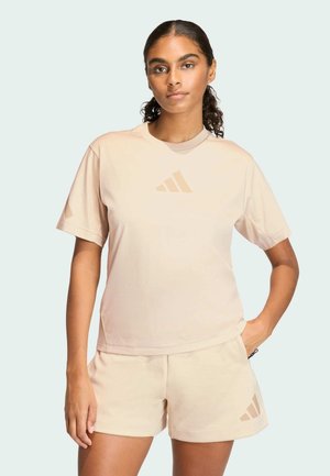Donna con capelli scuri raccolti in una coda di cavallo, indossa una t-shirt beige Adidas e pantaloncini, in piedi con una mano in tasca davanti a uno sfondo semplice.