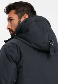 Dunkle Jacke mit Kapuze, die eine glatte Textur aufweist und ein kontrastierendes rotes Innenfutter hat. Metallische Akzente sind an der Kapuze sichtbar.