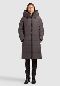 Sivi podstavljeni dugi kaput s velikim kapuljačom, zatvaranjem na zip i bočnim džepovima. Materijal ima glatku teksturu; nosi se preko crnih tajica i čizama.