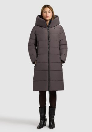 Grå vadderad långjacka med stor huva, dragkedjestängning och sidofickor. Materialet har en slät yta; bärs över svarta leggings och stövlar.