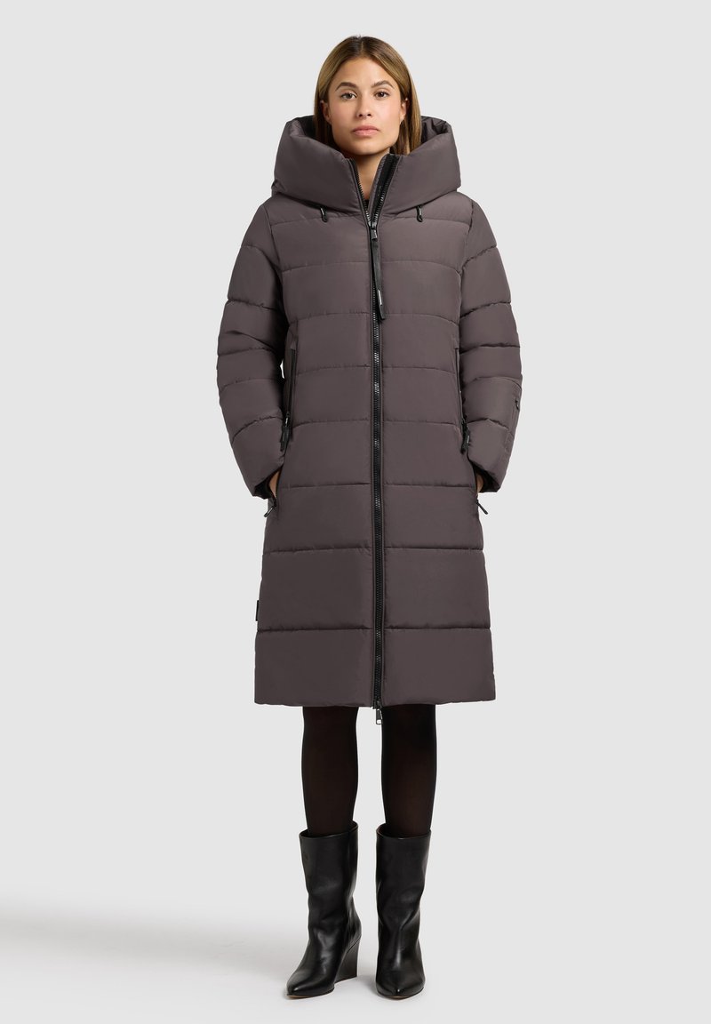 Grå polstret lang jakke med en stor hætte, gennemgående lynlås og sidelommer. Materialet har en glat tekstur; båret over sorte tights og støvler.