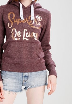 Osoba w bordowej bluzie z kapturem z napisem "Superdry De Luxe" w złotym kolorze i białymi sznurkami, ubrana w jasnoniebieskie, postrzępione szorty jeansowe.
