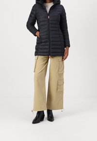 Černá prošívaná bunda s kapucí, zapínáním na zip a oranžovými doplňky, kombinovaná se světlými béžovými kalhotami typu cargo s několika kapsami a zapínáními.