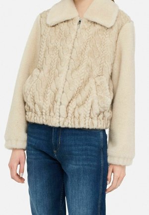 Femme portant une veste polaire beige texturée avec fermeture éclair et col, associée à un jean bleu raccourci aux hanches.