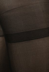 Schwarze, transparente Strumpfhosen mit glatter Textur, ausgestattet mit einem breiten, dunklen Taillenbund und einem dezenten Denier-Muster vom Knöchel bis zum Oberschenkel.
