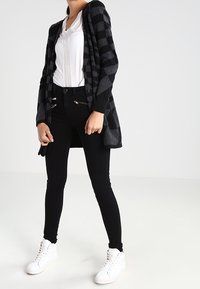 Cardigan a cuadros en negro y gris, hecho de tejido suave, combinado con una blusa blanca y jeans ajustados negros, llevado con zapatillas blancas.