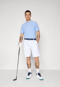 Világoskék, rövid ujjú póló CK logóval, fehér nadrág, sötétkék öv, fehér és kék golfcipő, golfütőt tart a kezében.