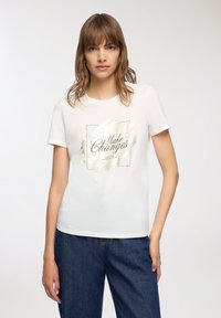 Biała bawełniana koszulka z krótkim rękawem, z nadrukiem ze złotej folii mówiącym "Wprowadzaj zmiany" na teksturowanym tle. Luźny krój, okrągły dekolt.