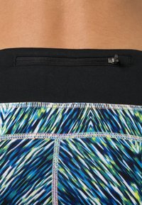 Gros plan sur une taille avec des leggings de sport bleus, verts et blancs à motifs, avec une ceinture noire et une petite poche zippée.