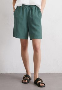 Shorts en lin verts avec une taille élastique et un cordon de serrage. Présentent deux poches latérales. Associés à des sandales noires à enfiler avec une boucle.