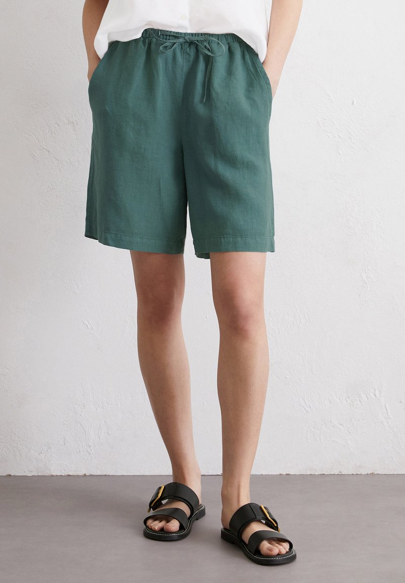 Shorts en lin verts avec une taille élastique et un cordon de serrage. Présentent deux poches latérales. Associés à des sandales noires à enfiler avec une boucle.