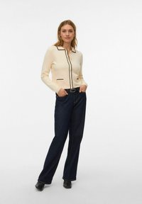 Kremowy kardigan z czarnym wykończeniem, zapinany na guziki, z dwoma przednimi kieszeniami, w połączeniu z ciemnoniebieskimi szerokimi dżinsami i czarnym obuwiem.