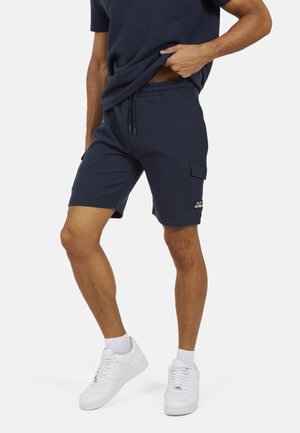 Uomo che indossa pantaloncini cargo blu navy con trama, sneakers bianche e una maglietta coordinata blu navy, solleva leggermente la maglietta per mostrare la vita dei pantaloni.
