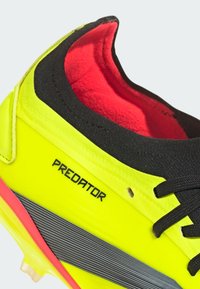 Chaussure de football jaune vif avec des accents noirs, dessus texturé, doublure rouge, rayures grises et marquage "PREDATOR" sur le côté.