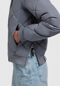 Grijze geborduurde jas met een soepele stof, elastische manchetten en een ritssluiting aan de voorkant. Lichte blauwe denim rok zichtbaar eronder.