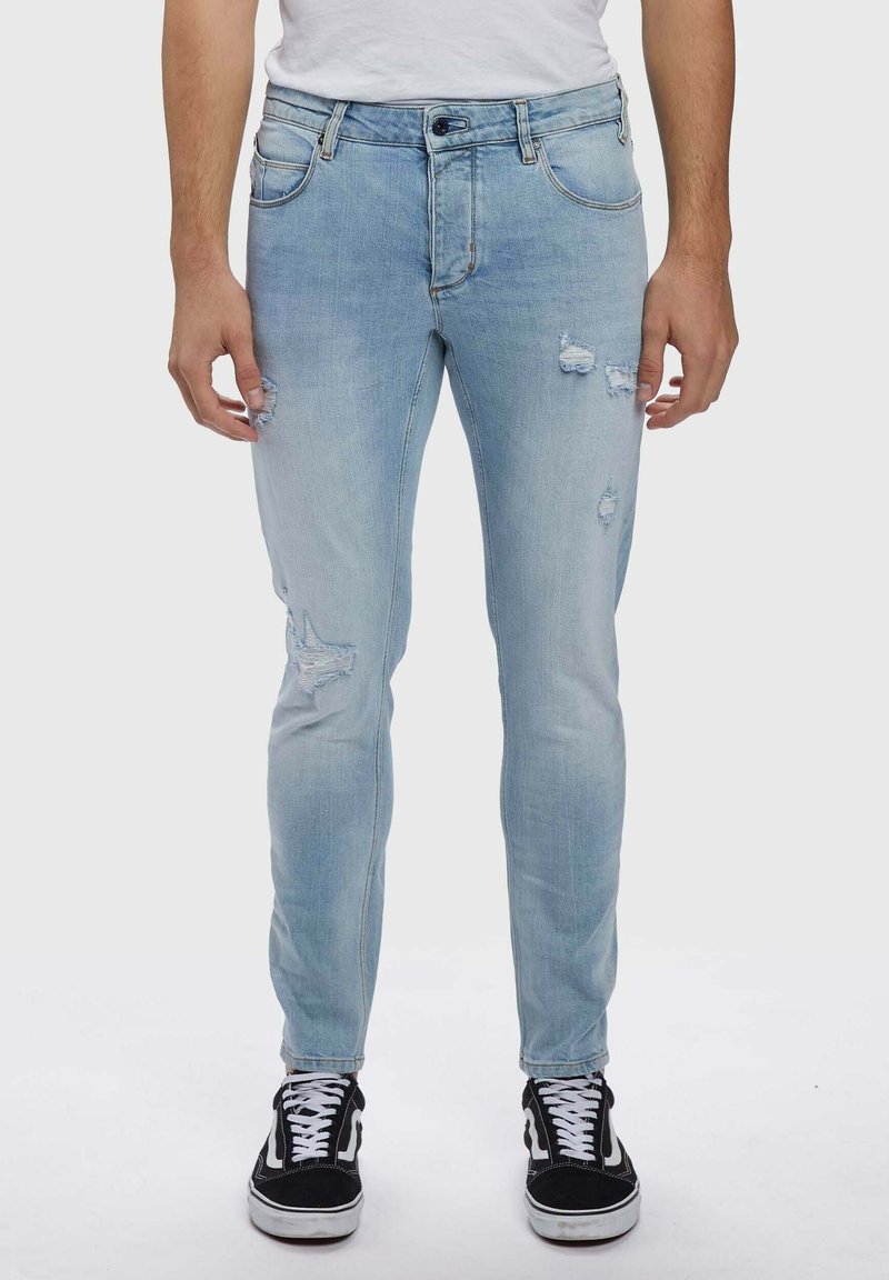 Světlé modré džíny z denimu s úzkým střihem, s efektu opotřebení a třepícími se detaily. Nošeny s černými teniskami.