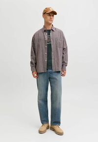 Camisa de cuadros en rojo, azul y blanco sobre una camiseta oscura, combinada con unos pantalones vaqueros de color azul claro y zapatos marrones, completada con una gorra de color marrón.