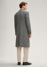 Cappotto in tweed grigio con vestibilità sagomata, dotato di spacco posteriore singolo, rever a incavo e dettagli neri, abbinato a pantaloni beige e scarpe nere.