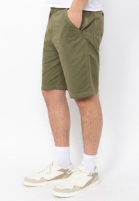Pantalones cortos de algodón verde oliva con bolsillos laterales, corte clásico, hasta la rodilla y dobladillo cosido. Combinados con zapatillas blancas y calcetines.