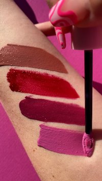 Fem färger av flytande läppstift på en ljus hudton-arm: brun, röd, mörk rosa, djup lila och klar rosa. Slät textur, glansig finish.