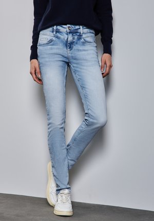 Jean bleu clair avec une coupe slim, design à cinq poches, passants de ceinture et décoloration subtile. Associé à des baskets blanches sur une surface grise.