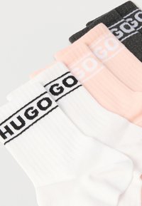 Štyri páry rebrovaných ponožiek v bielej, svetloružovej a tmavosivej farbe. Každý pár má čierny pruh s logom „HUGO“ blízko manžety.