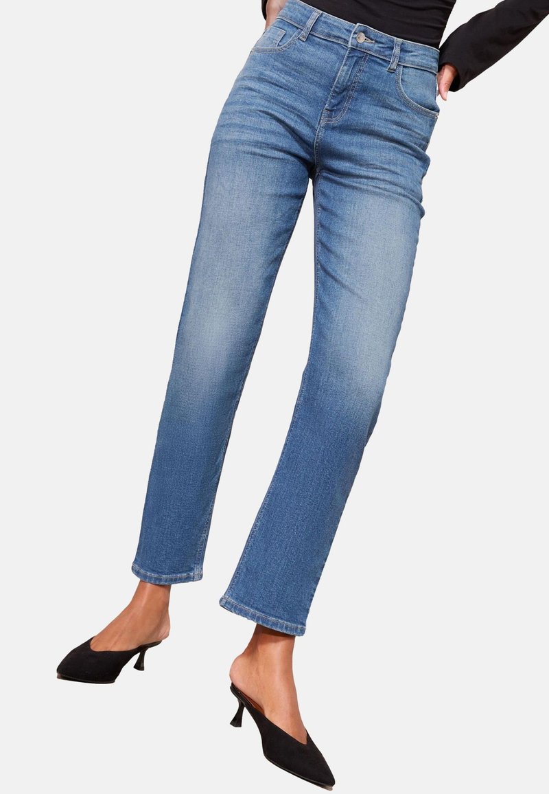Femme portant un jean bleu taille mi-haute, coupe droite, et des chaussures noires plates à enfiler, debout les mains dans les poches.