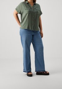Grøn kortærmet skjorte med knaplukning og to brystlommer, parret med lyseblå brede jeans og sorte sandaler med platåsåler.