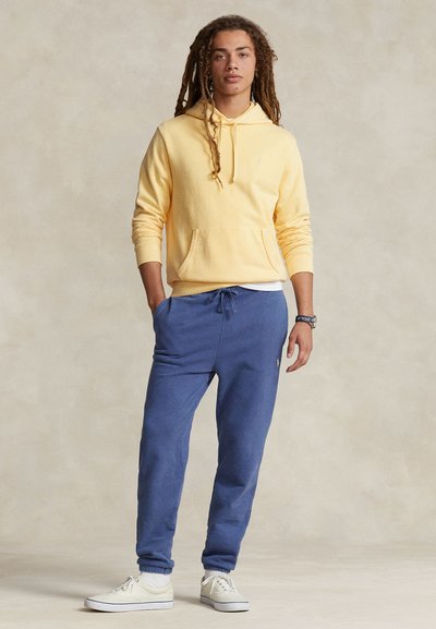 Κίτρινο φούτερ με κουκούλα και μπροστινή τσέπη, συνδυασμένο με μπλε τζόγκερ. Λευκό στρωμένο πουκάμισο από κάτω. Το μοντέλο φοράει λευκά αθλητικά παπούτσια και στέκεται σε ουδέτερο φόντο.
