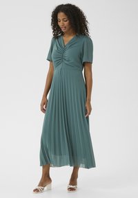 Teal pleated dress with short sleeves, V-neckline, and gathered bodice, paired with white strappy sandals on a plain background.