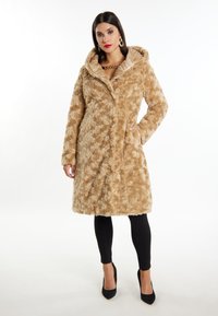 Beige műszőr kabát kapucnival, texturált felülettel, hosszú ujjak, elülső gombos záródás és oldalsó zsebek. Fekete leggings és magassarkú felett viselve.