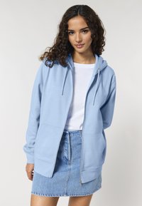 Felpa con zip di colore azzurro chiaro, dotata di cappuccio, tasche frontali e hardware in metallo argentato, abbinata a una t-shirt bianca e a una gonna di denim.