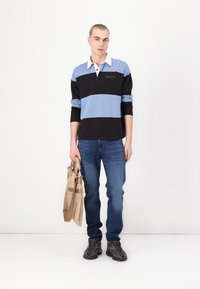 Črno-modra polo majica s belim ovratnikom, modri in črni jeans, drži svetlo rjavo torbo. Robustni črni športni copati dopolnjujejo obleko.