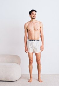 Beige boxershorts met een zwarte elastische tailleband. Glad materiaal, knoopsluiting aan de voorkant, relaxte pasvorm. Model staat blootsvoets tegen een lichte achtergrond.
