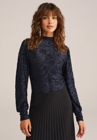 Marinblå blommönstrad långärmad topp med texturerad tyg och hög krage, tillsammans med en svart plisserad kjol.