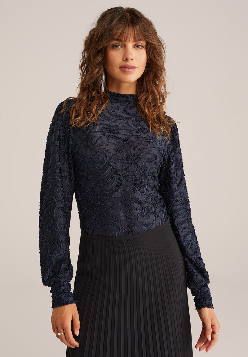 Marinblå blommönstrad långärmad topp med texturerad tyg och hög krage, tillsammans med en svart plisserad kjol.